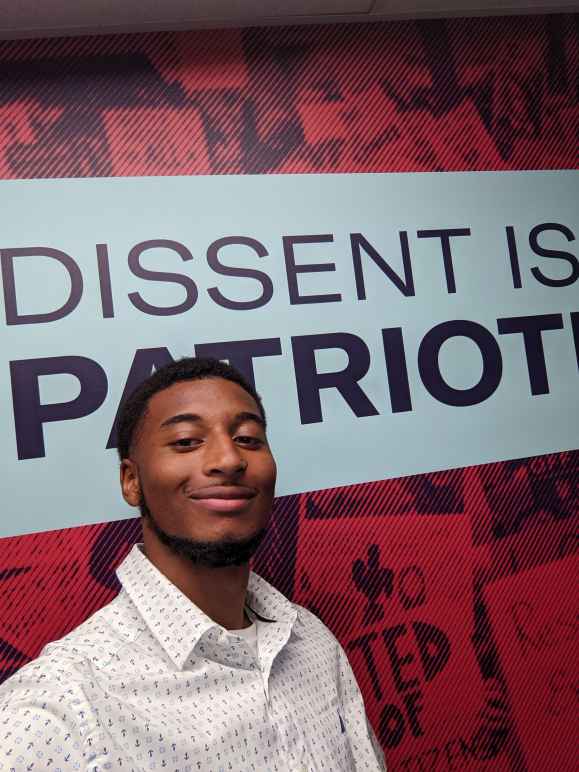 A Black man with short hair and a beard wearing a white button up shirt with a light blue pattern standing in front of a sign that reads Dissent is Patriotic